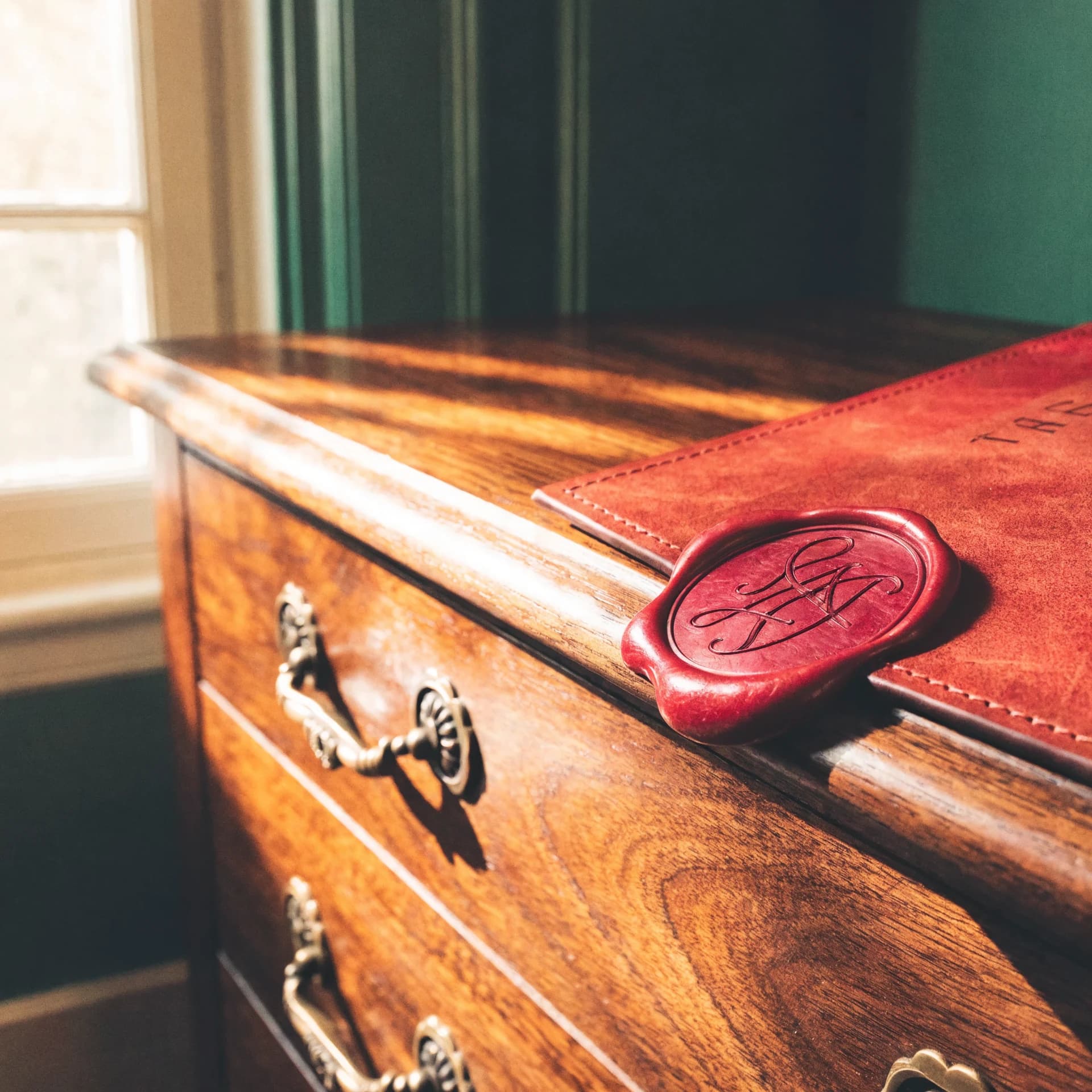 A leather-bound thank-you card with a deep garnet wax seal pressed with a monogram, on a polished walnut chest.