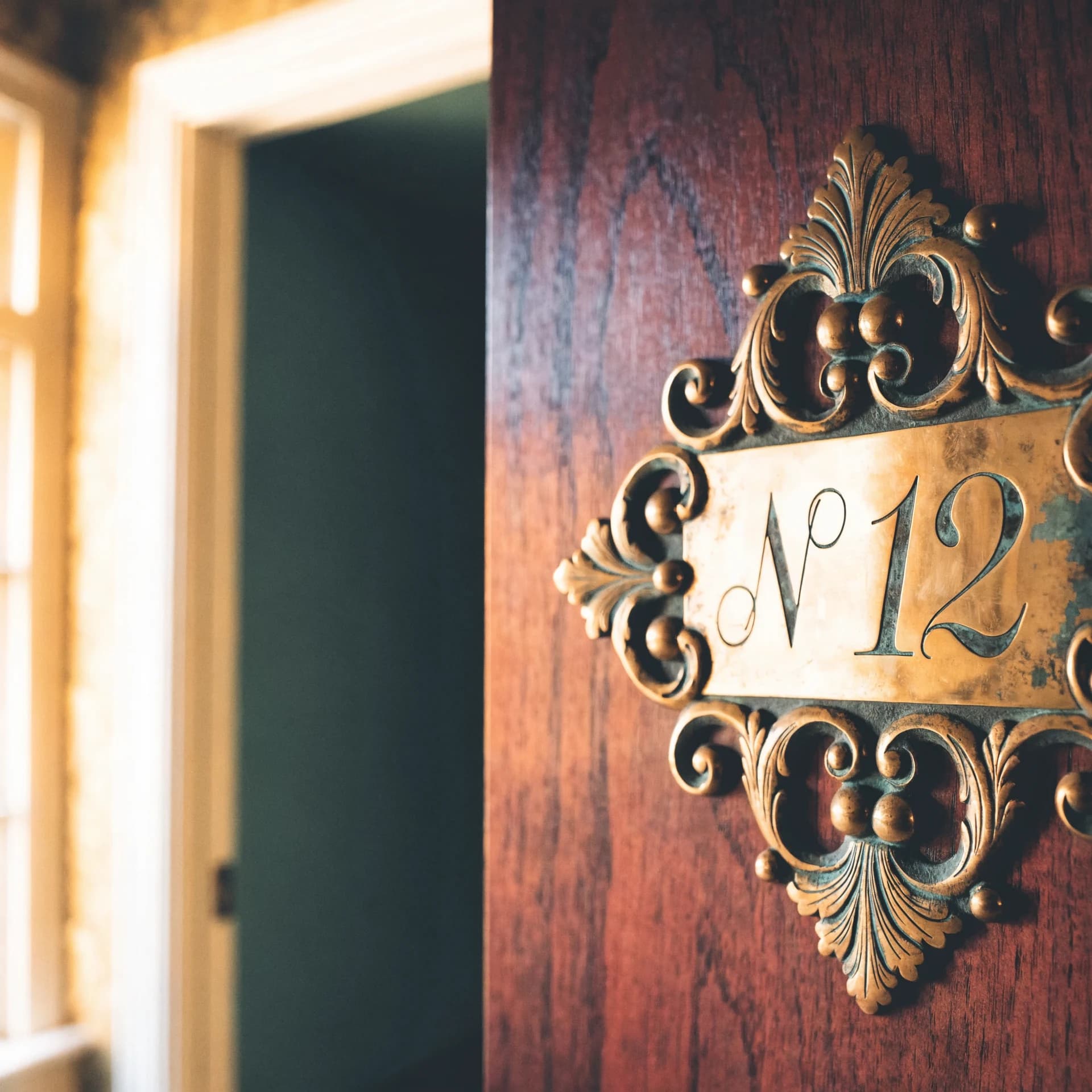 An engraved brass room-number plate reading 'No 12' set in a Victorian-scroll brass frame on a stained walnut hotel door.